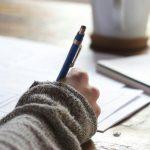 Person Writing on Brown Wooden Table Near White Ceramic Mug